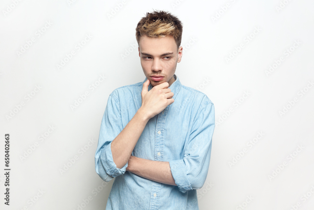 Portrait of pensive thoughtful handsome man wearing denim shirt standing holding his chin, being deep in thoughts, thinking about his work. Indoor studio shot isolated on gray background. Portrait of pensive thoughtful handsome man wearing denim shirt standing holding his chin, being deep in thoughts, thinking about his work. Indoor studio shot isolated on gray background.