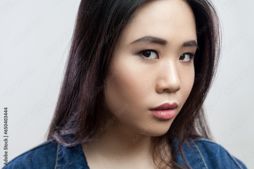 Naklejka premium Closeup portrait of serious concentrated beautiful young adult brunette woman with makeup and long hair, looking at camera. Indoor studio shot isolated on gray background.