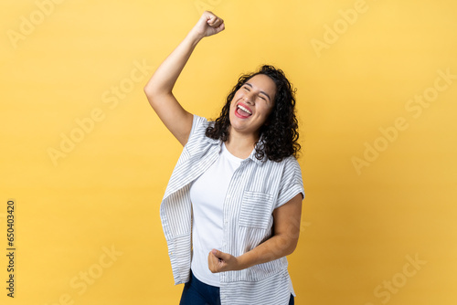 Portrait of excited happy woman with dark wavy hair expressing winning gesture with raised fists and screaming, celebrating victory. Indoor studio shot isolated on yellow background.