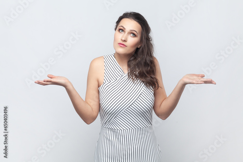 Portrait of brunette woman spreads palms, shrugs shoulders with perplexed expression, being indecisive, has no idea what happened, wearing striped dress. Indoor studio shot isolated on gray background photo