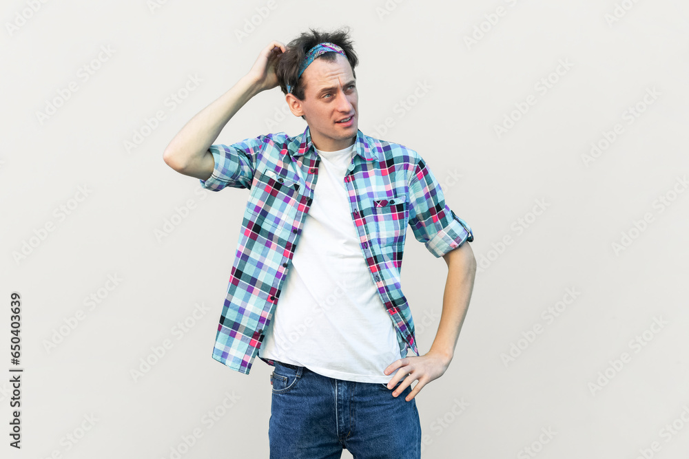 Indignant puzzled man rubbing head, tries to remember necessary information, looks in displeasure, wearing blue checkered shirt and headband. Indoor studio shot isolated on gray background. Indignant puzzled man rubbing head, tries to remember necessary information, looks in displeasure, wearing blue checkered shirt and headband. Indoor studio shot isolated on gray background.