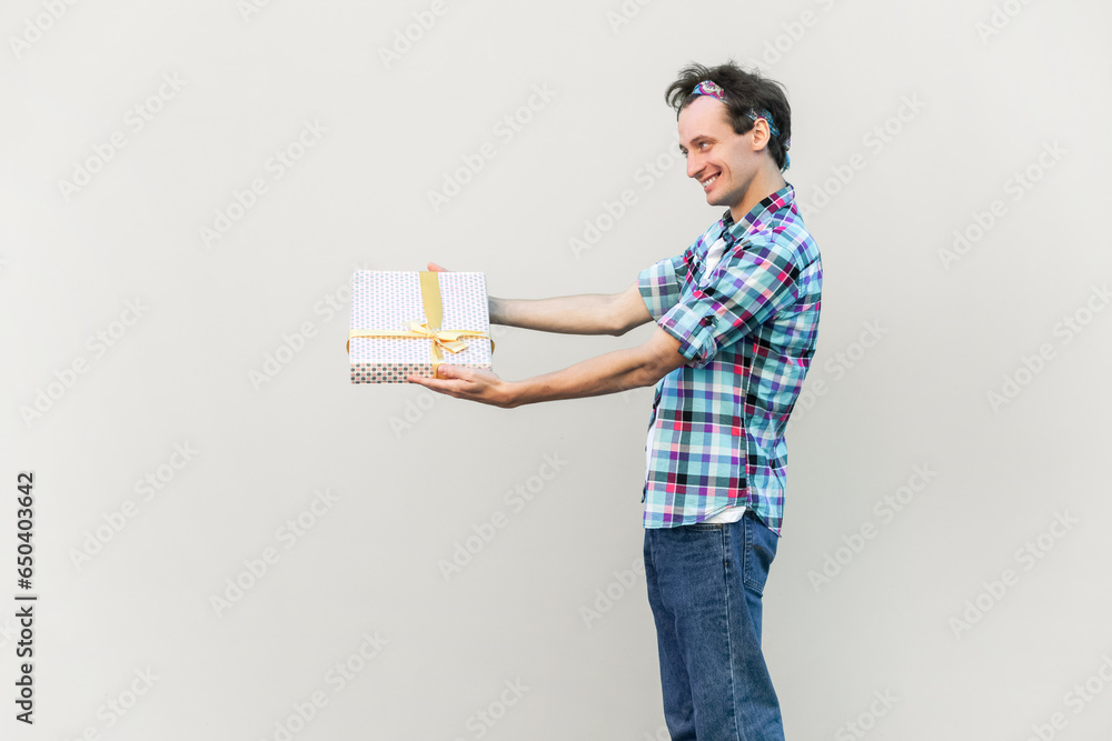Side view of positive happy smiling standing with outstretched hands, giving present to his girlfriend, wearing blue checkered shirt and headband. Indoor studio shot isolated on gray background. Side view of positive happy smiling standing with outstretched hands, giving present to his girlfriend, wearing blue checkered shirt and headband. Indoor studio shot isolated on gray background.