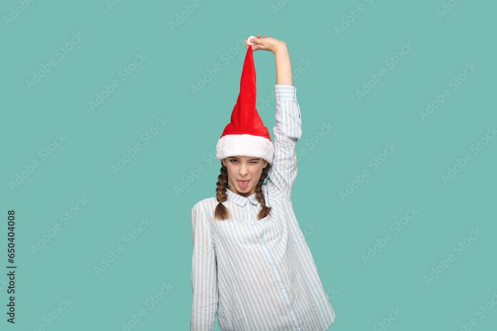 Φωτογραφία Portrait of happy funny teenager girl with braids wearing striped shirt, looking at camera with tongue out, pulling up her Santa Claus hat