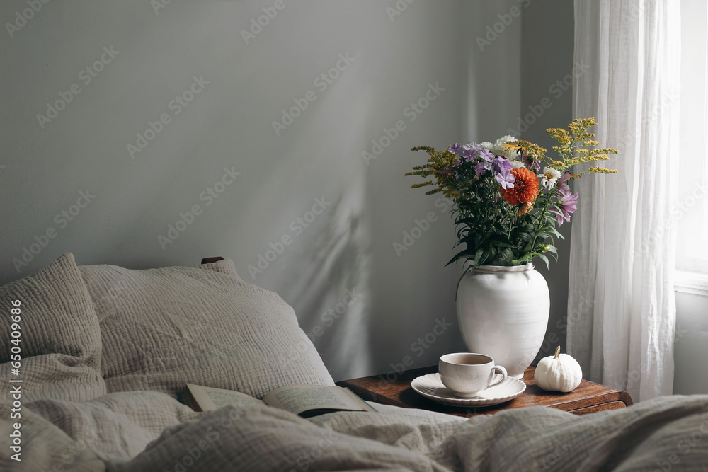 Elegant moody bedroom interior. Cup of coffee, pumpkin on retro wooden bedside table. White ceramic vase. Bouquet of dahlia, cosmos, solidago flowers. Beige muslin cushions, book in bed near window. - obrazy, fototapety, plakaty 