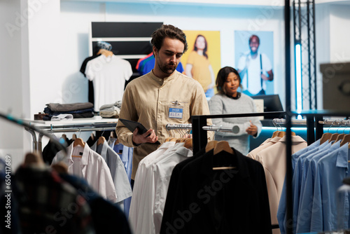 Clothing store employee examining apparel rack and displayed merchandise while using digital tablet. Shopping mall boutique worker doing inventory management with portable gadget