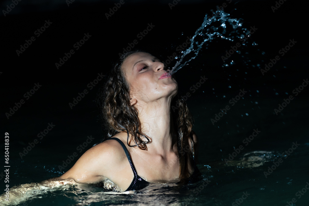 Obraz premium Curly beauty in the sea at night splashing water from her mouth wearing a blue swimsuit