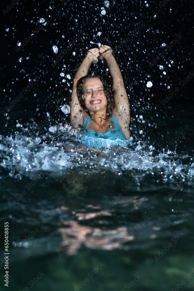 Obraz premium A beautiful and curly young woman in a blue swimsuit in the sea at night splashing water everywhere