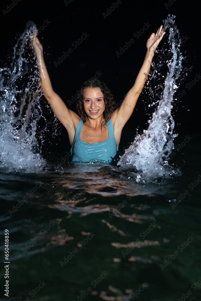 A beautiful and curly young woman in a blue swimsuit in the sea at night splashing water in the shape of wings A beautiful and curly young woman in a blue swimsuit in the sea at night splashing water in the shape of wings