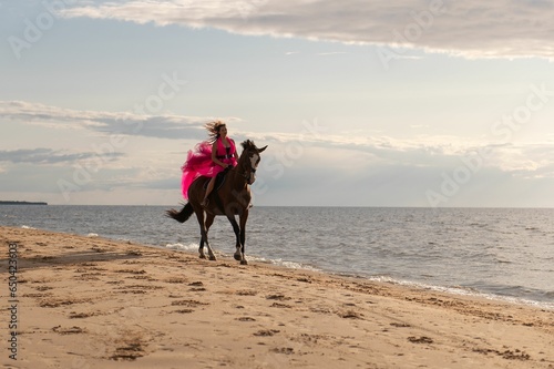 Young woman wearing a vibrant pink dress riding a horse on a beach