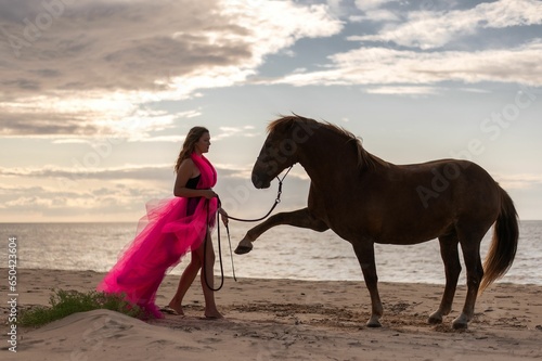 Young woman wearing a vibrant pink dress posing with a horse on a beach