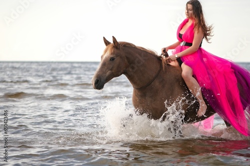 Young woman wearing a vibrant pink dress riding a horse in the water