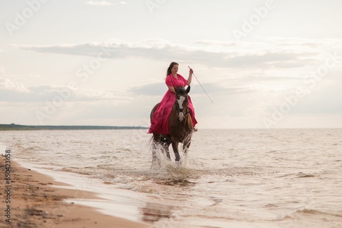 Beautiful young female in a bright pink dress riding a horse on a beach