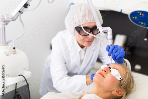 Female client receiving face skin laser resurfacing procedure from professional beautician on modern equipment in clinic of aesthetic cosmetology