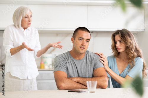 Young couple quarrel with mother while sitting at table in kitchen