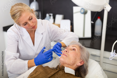 Experienced female cosmetologist working with senior female client in clinic of aesthetic medicine, injecting facial filler into skin to correct face contour