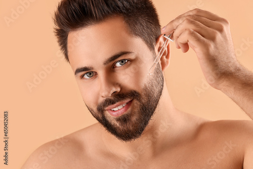 Handsome man applying cosmetic serum onto face on beige background, closeup
