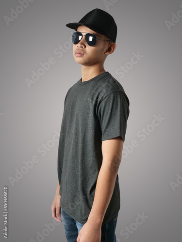 Young Asian teenage boy wearing black shirt, hat and sunglasses standing with hands on hip over grey background, t-shirt template