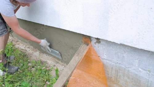 A worker uses a large trowel to putty the foundation of a house. Covering the foundation with mesh and putty. A man plasters a house and insulates it