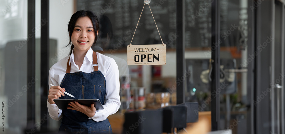 Fototapeta premium Successful small business owner in casual wearing jeans apron holding tablet standing at entrance