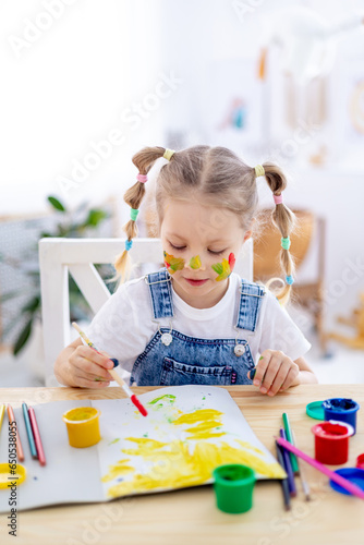a little child girl paints a drawing in an album and smiles with a dirty face and hands, a happy child draws a picture at home at the table