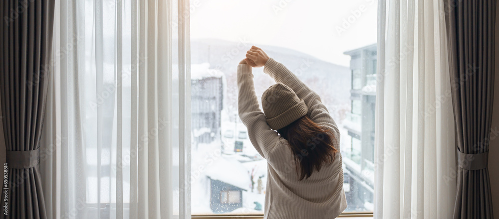 young woman in sweater looking through the window in winter season, happy female rising arms and stretching after waking at apartment or home in the morning. Lifestyle and Relaxing concepts young woman in sweater looking through the window in winter season, happy female rising arms and stretching after waking at apartment or home in the morning. Lifestyle and Relaxing concepts