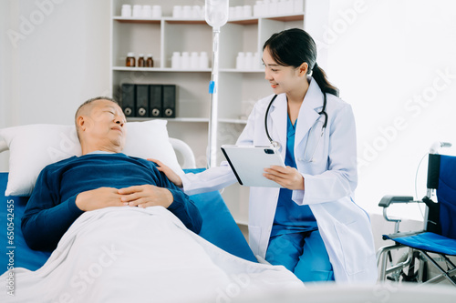 Asian doctor in white suit take notes while discussing and Asian elderly, man patient who lying on bed with receiving saline solution in hospital .