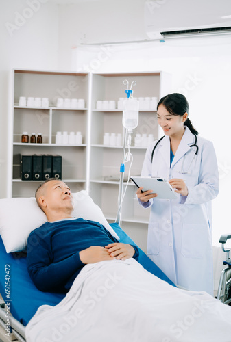 Asian doctor in white suit take notes while discussing and Asian elderly, man patient who lying on bed with receiving saline solution in hospital .