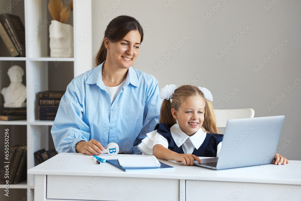 Young mother sits at the table with her daughter and helps a little schoolgirl in uniform do her homework on the computer at home. A woman tutor teaches a girl lessons on a laptop Young mother sits at the table with her daughter and helps a little schoolgirl in uniform do her homework on the computer at home. A woman tutor teaches a girl lessons on a laptop