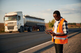 In orange colored uniform, with tablet. Black man is standing on the road with truck on it