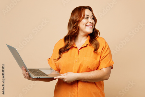 Young ginger chubby overweight IT woman wears orange shirt casual clothes hold use work on laptop pc computer look aside isolated on plain pastel beige background studio portrait. Lifestyle concept. photo