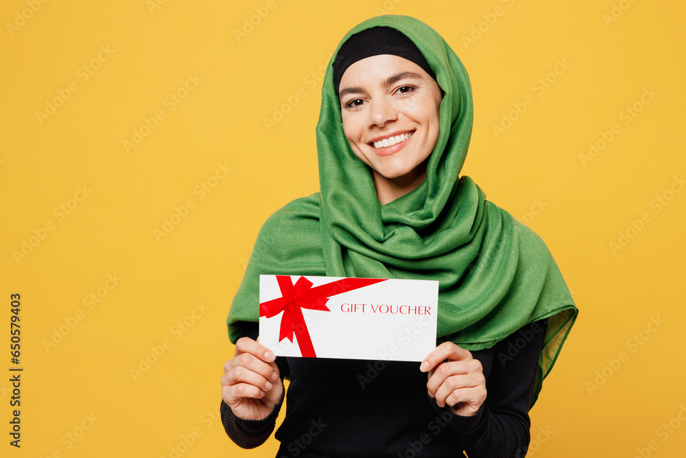 Young asian muslim woman wear green hijab abaya black clothes hold gift certificate coupon voucher card for store isolated on plain yellow background People uae middle eastern islam religious concept Young asian muslim woman wear green hijab abaya black clothes hold gift certificate coupon voucher card for store isolated on plain yellow background People uae middle eastern islam religious concept