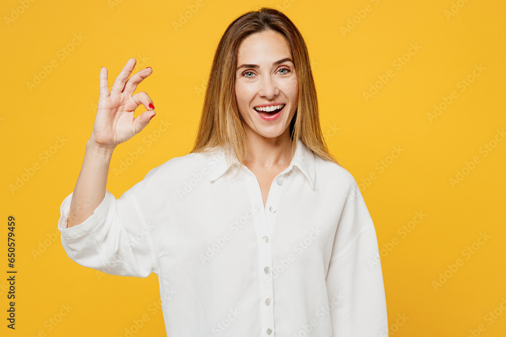 Young smiling satisfied positive fun caucasian happy woman she wears white shirt casual clothes look camera show ok okay gesture isolated on plain yellow background studio portrait. Lifestyle concept. Young smiling satisfied positive fun caucasian happy woman she wears white shirt casual clothes look camera show ok okay gesture isolated on plain yellow background studio portrait. Lifestyle concept.