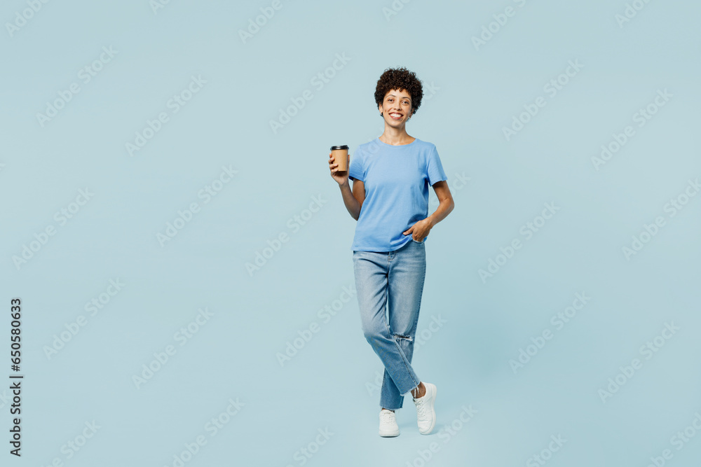 Full body smiling young woman of African American ethnicity wear t-shirt casual clothes hold takeaway delivery craft paper brown cup coffee to go isolated on plain pastel light blue cyan background. .