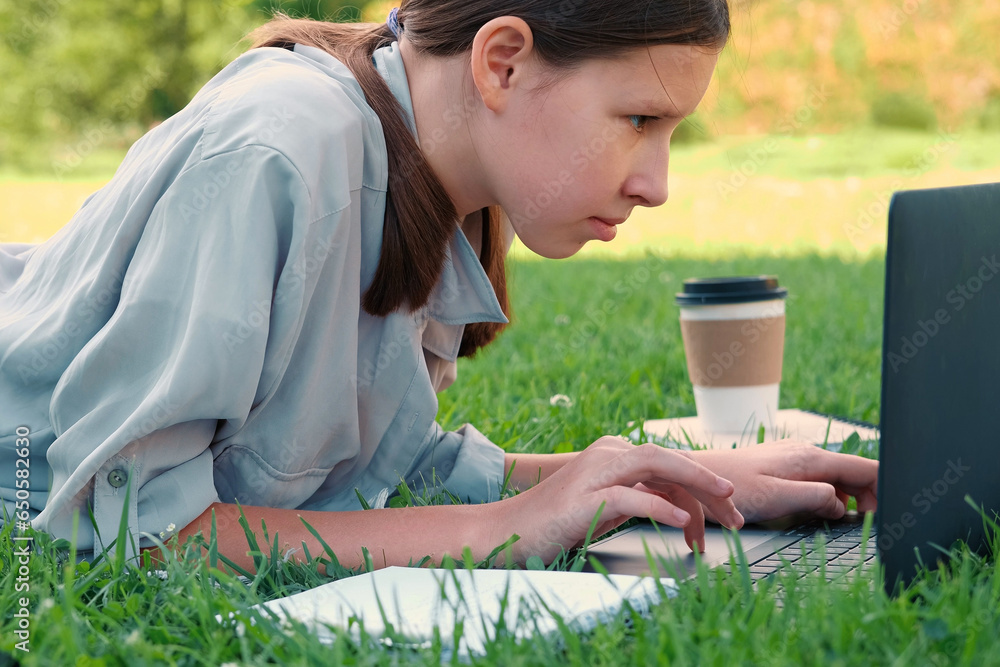 Teenage schoolgirl studying reading her books, tablet and notebook, sitting outdoors. Back to school. Student girl lying on the green grass using laptop in the college yard or park. Distance learning. Teenage schoolgirl studying reading her books, tablet and notebook, sitting outdoors. Back to school. Student girl lying on the green grass using laptop in the college yard or park. Distance learning.