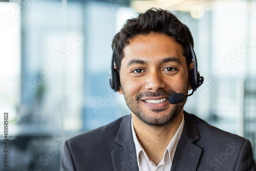 Close-up portrait of young successful businessman with headset phone, customer support worker in business suit smiling and looking at camera, workplace inside office.