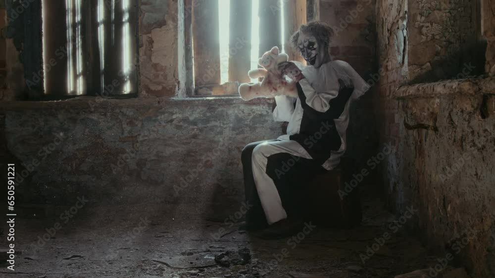 Long shot of clown psychopath sitting alone in abandoned place with shabby walls and floor, playing with toy bear Long shot of clown psychopath sitting alone in abandoned place with shabby walls and floor, playing with toy bear