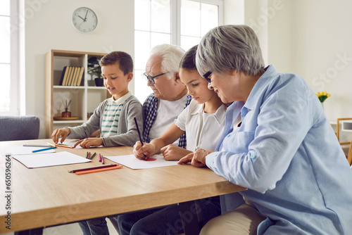 Senior couple grandparents spending time with their grandchildren boy and girl at home on weekend drawing with colored pencils together sitting at the table. Family leisure concept.