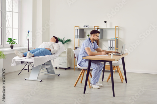 Fat overweight woman lying on the couch in clinic receiving IV drip infusion or vitamin therapy in blood with a doctor working at the desk in office. Sick plus size girl receiving injection therapy.