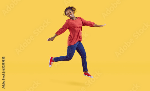 Happy young black woman having lots of fun in the studio. Cheerful carefree African American girl wearing comfortable blue jeans jumping high in the air isolated on a vibrant yellow colour background