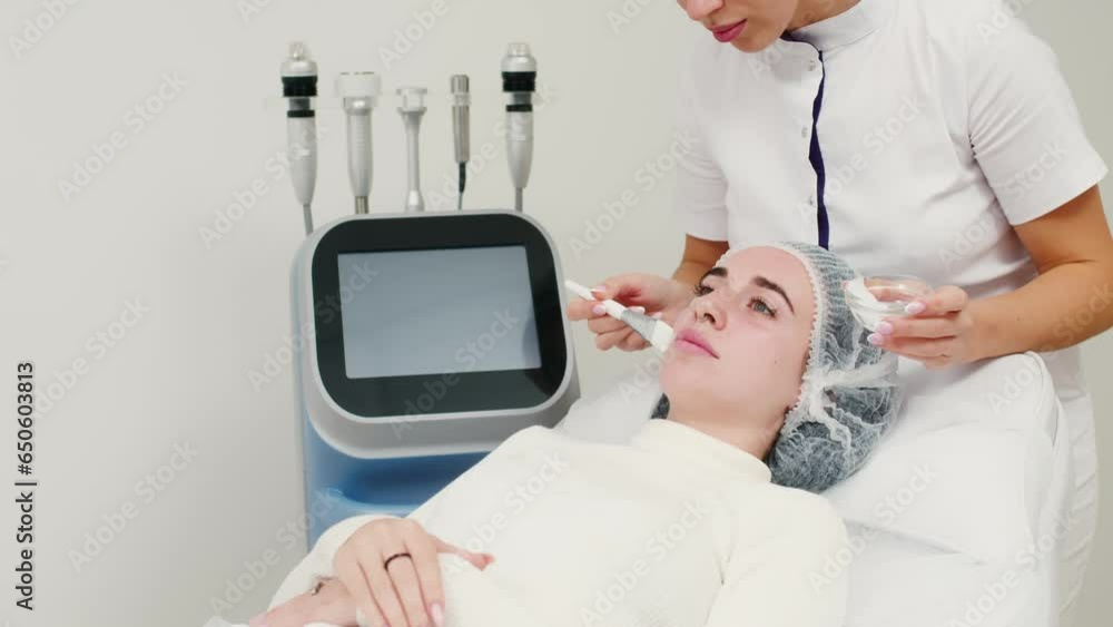 A cosmetologist applies anesthetic cream to a young woman's face before a facial cosmetic procedure. Anesthetic cream. Preparation for healing, tightening, skin rejuvenation in beauty salons. A cosmetologist applies anesthetic cream to a young woman's face before a facial cosmetic procedure. Anesthetic cream. Preparation for healing, tightening, skin rejuvenation in beauty salons.