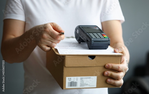Courier holds cardboard box with payment terminal and documents with pen. Parcel delivery and electronic payment concept