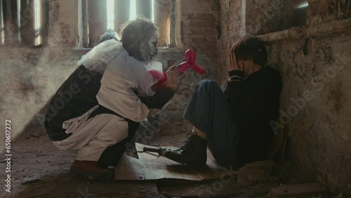 Long shot of clown psycho in white clothes scaring hostage woman with balloon toy in creepy place