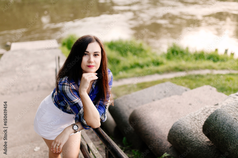 A girl with long dark hair, dressed in a checkered blue shirt, a white skirt, poses against the background of a river and stone steps.Portrait of a young beautiful woman against a background of nature A girl with long dark hair, dressed in a checkered blue shirt, a white skirt, poses against the background of a river and stone steps.Portrait of a young beautiful woman against a background of nature