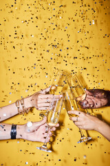 Closeup of group of people clinking champagne glasses against vibrant yellow background with confetti, copy space