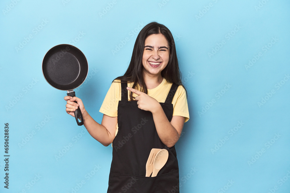 Obraz premium Young woman with pan on blue studio smiling and pointing aside, showing something at blank space.