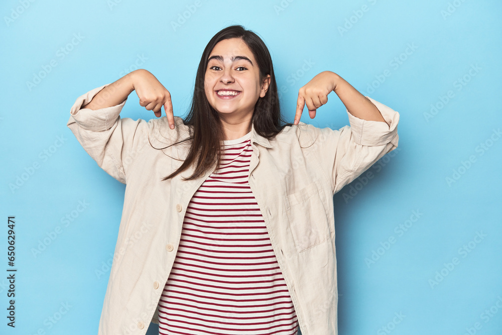 Young Caucasian woman on blue backdrop points down with fingers, positive feeling. Young Caucasian woman on blue backdrop points down with fingers, positive feeling.