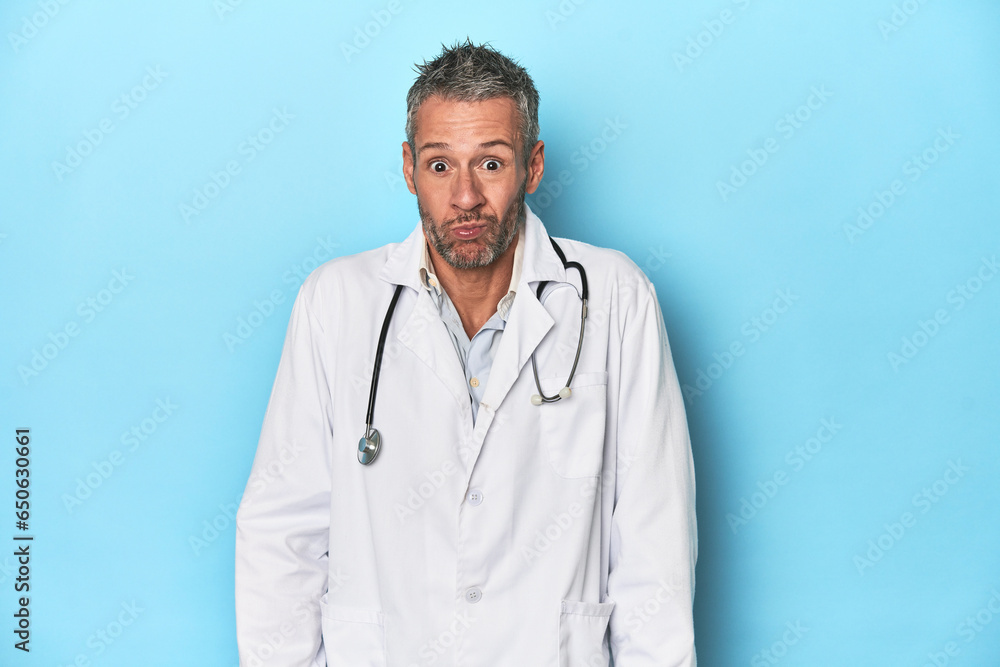 Caucasian middle-aged doctor on blue background shrugs shoulders and open eyes confused. Caucasian middle-aged doctor on blue background shrugs shoulders and open eyes confused.