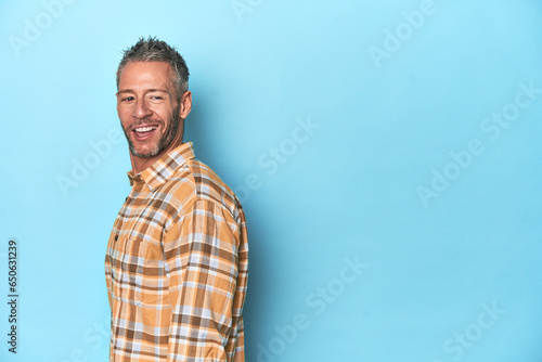 Middle-aged caucasian man on blue backdrop looks aside smiling, cheerful and pleasant.