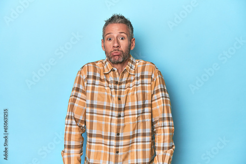 Middle-aged caucasian man on blue backdrop shrugs shoulders and open eyes confused.