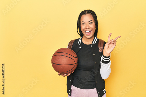 Indonesian schoolgirl with basketball on yellow joyful and carefree showing a peace symbol with fingers.